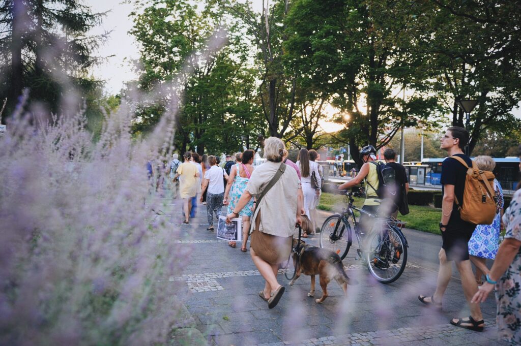 Kolorowa, pozioma fotografia: szerokim chodnikiem idzie duża grupa osób w różnym wieku. Ktoś idzie z psem, inna osoba prowadzi rower. Trwa spacer z oprowadzaniem po osiedlu. Po lewej stronie zdjęcia znajdują się kwitnące na fioletowo rośliny.