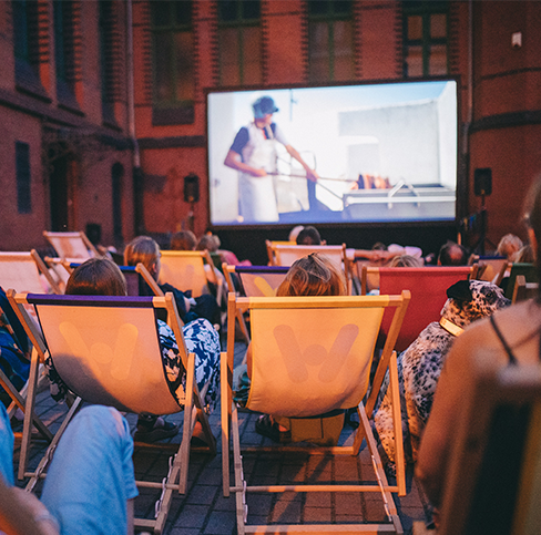 Кольорова фотографія, зроблена під час кінопоказу просто неба. Група людей різного віку сидить на шезлонгах, перед ними встановлено великий екран.