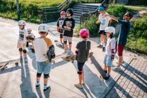 fotografia przedstawia grupę dzieci z deskorolkami zebranych w kole na skateparku, instruktor coś im tłumaczy