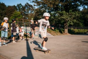 fotografia przedstawia chłopca jadącego na deskorolce po skateparku, w tle widoczna grupa osób z deskorolkami, czekająca na swoją kolej przejazdu.
