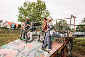 fotografia przedstawia grupę dzieci w różnym wieku z hulajnogami stojącą na przeszkodzie na skateparku