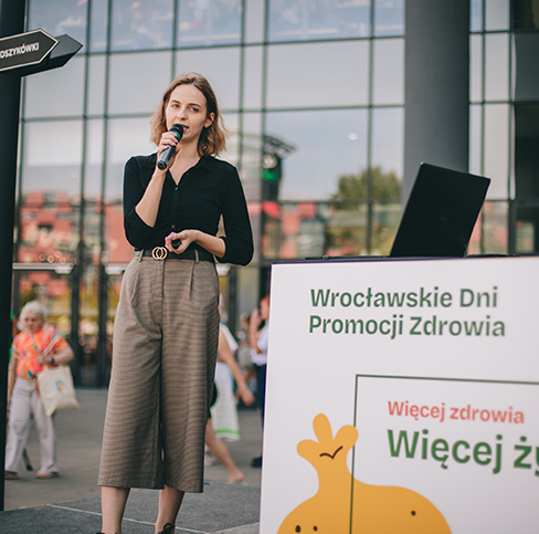 Кольорова фотографія, зроблена під час минулорічного заходу. Молода жінка стоїть на сцені перед торговим центром «Магнолія» та говорить у мікрофон.