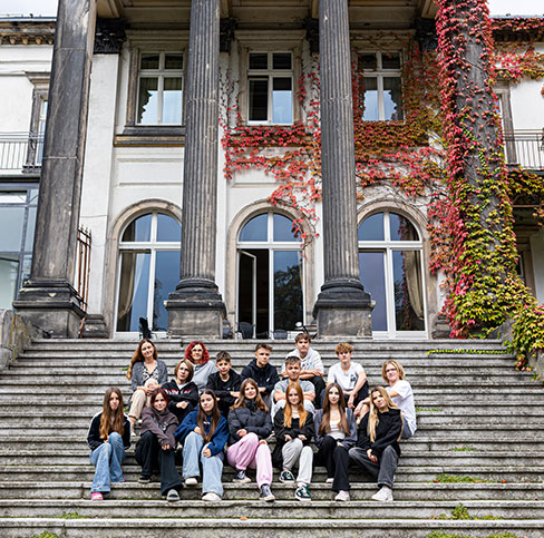 Kolorowa fotografia przedstawia grupę osób siedzących na schodach prowadzących do pałacu; w tle widać pałac i kolumny częściowo porośnięte bluszczem