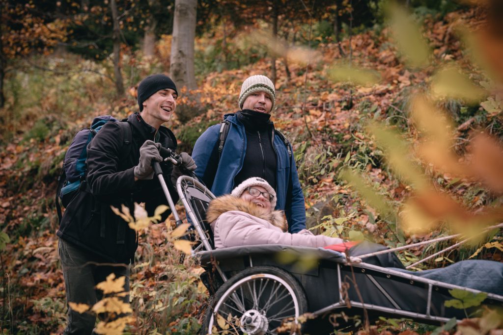 Kolorowa fotografia przedstawia młodego mężczyznę pchającego trójkołowy wózek po lesie. W wózku siedzi młoda, roześmiana kobieta. Obok idzie drugi mężczyzna.