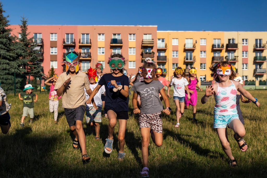 Kolorowa fotografia wykonana podczas wakacyjnych zajęć dla dzieci. Grupa dzieci w kolorowych maskach z papieru biegnie w kierunku fotografki.