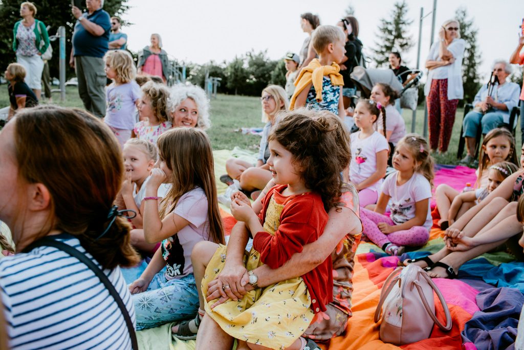 Kolorowa fotografia wykonana podczas plenerowego występu teatrzyku dla dzieci. Na łące siedzi duża grupa dzieci wraz z rodzicami. Wszyscy w skupieniu oglądają występ.
