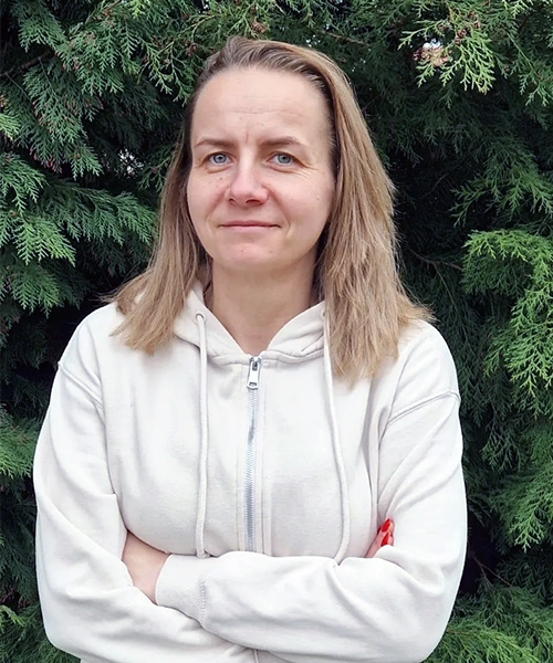 На кольоровій фотографії зображена молода жінка на ім'я Кася. У Касі світле волосся до плечей і біла блузка. Позаду неї видно дерева.