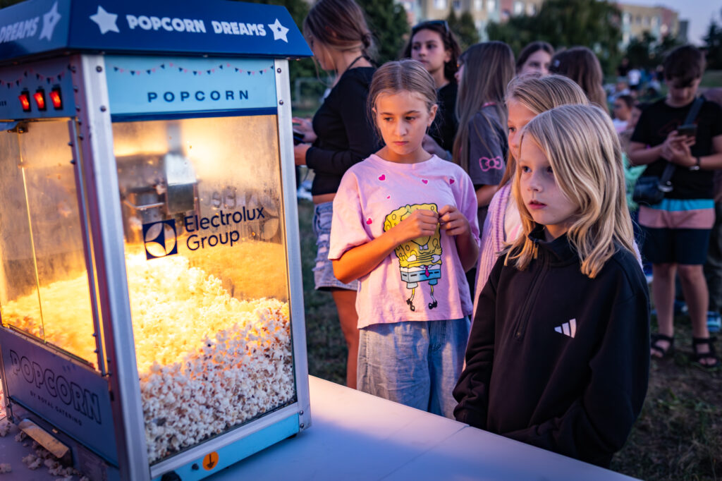 Children and teenagers queue for popcorn during an outdoor event. To the left is an illuminated popcorn machine with the Electrolux Group logo, and girls are waiting at it. In the background are other participants.