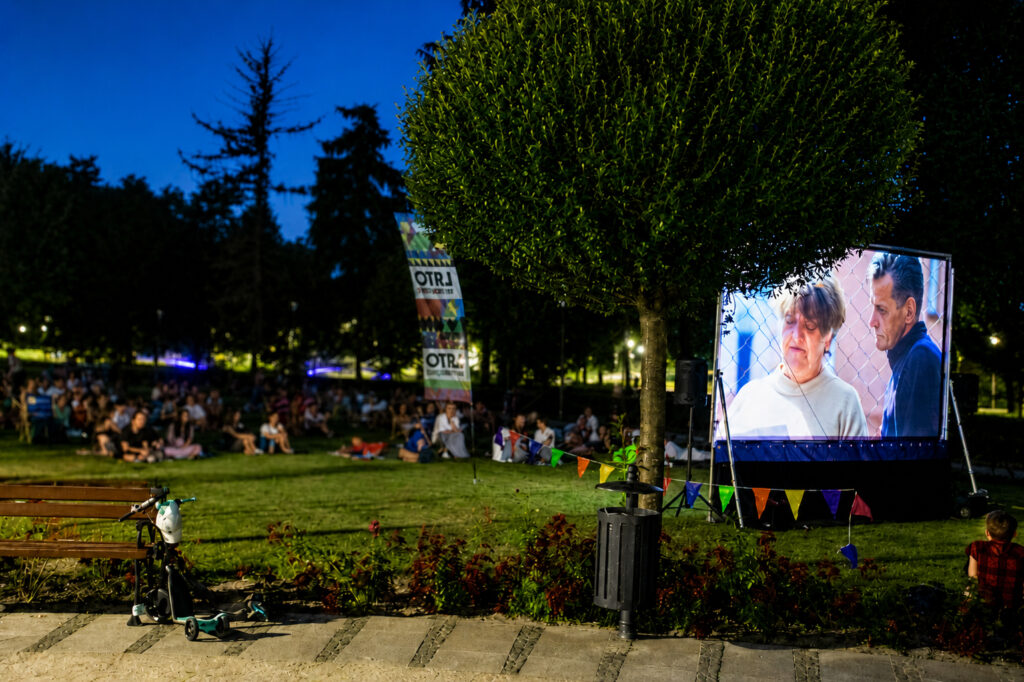 An evening outdoor cinema screening in the park. The audience sits on the lawn, watching a film projected on a large screen. Trees surround the area, and lamps illuminate the space. In the foreground, benches, a scooter, and colorful pennants hang near the screen.